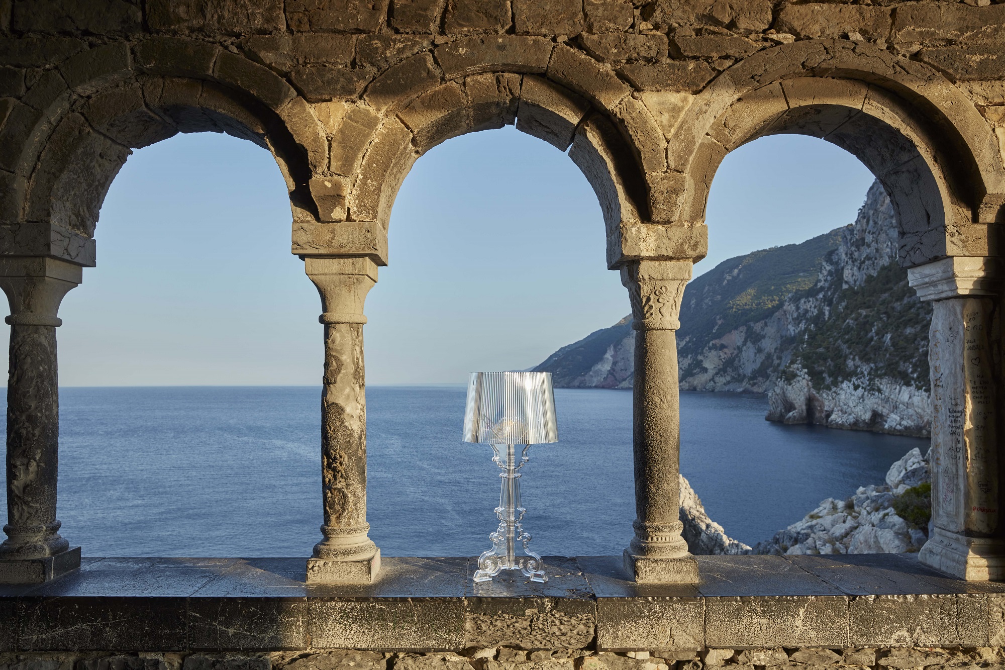 Bourgie Tischleuchte von Kartell, glasklar und dimmbar, vor malerischer Küstenlandschaft mit Meer und Felsen.