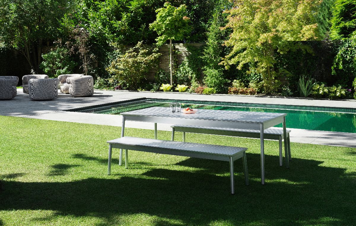 Normann Copenhagen Ex Tisch Outdoor mit Bänken auf Rasen vor Pool und üppigem Grün.