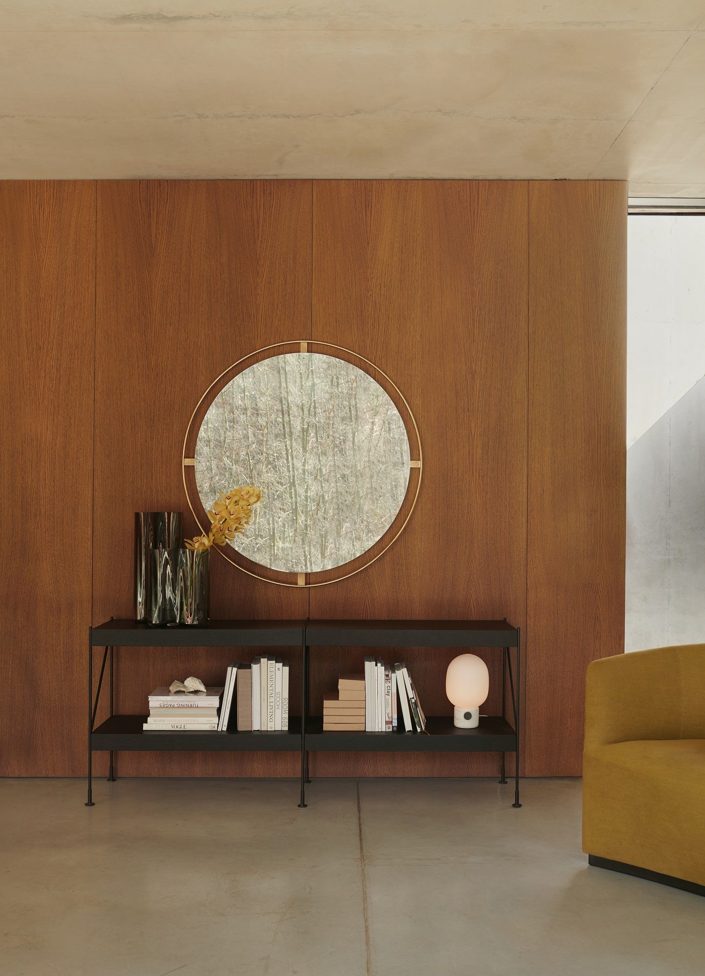 Audo Copenhagen Nimbus Mirror über einem schwarzen Regal mit Büchern und Dekoration vor Holzpaneelen.