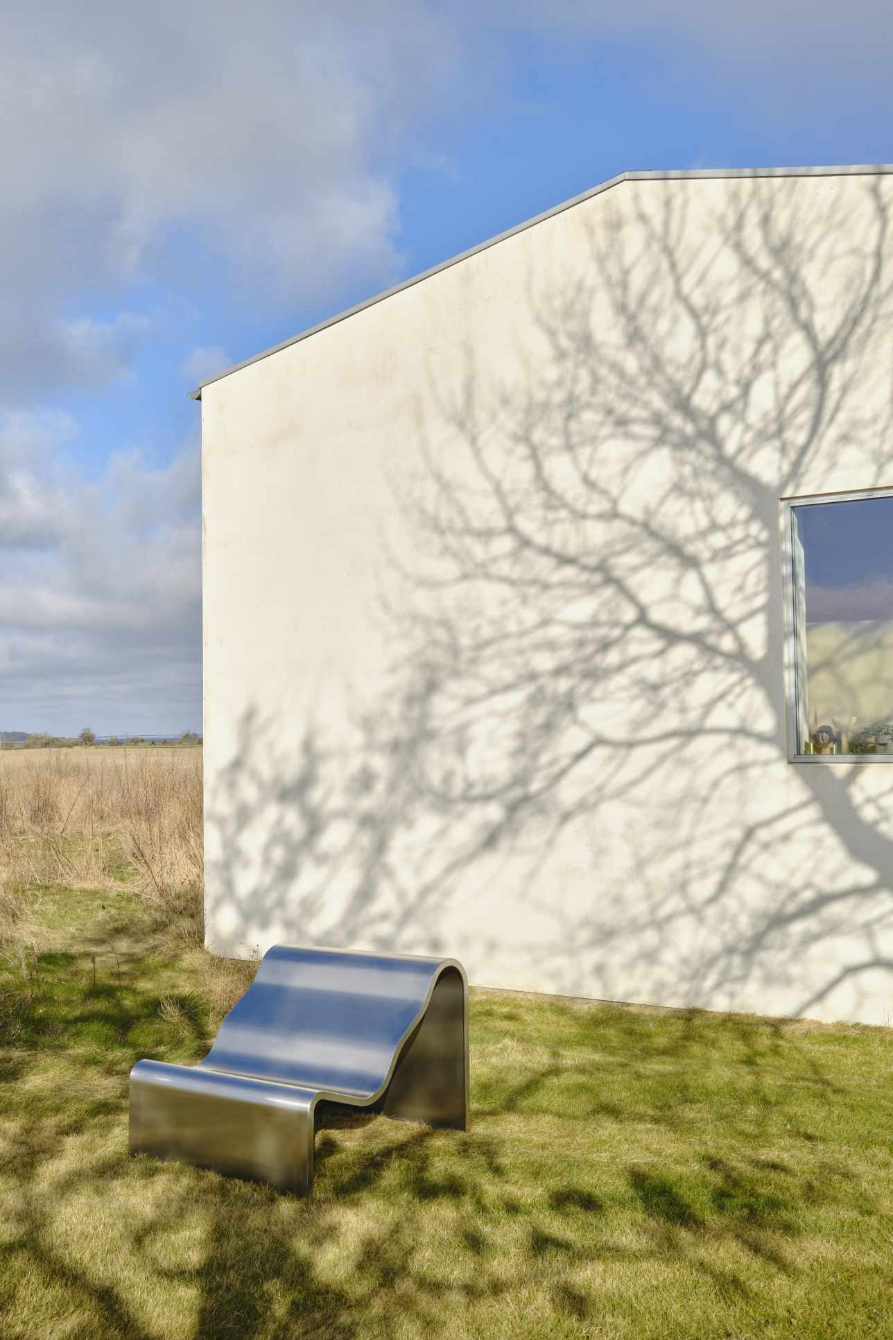 Muuto Dream Bench Outdoor Bank aus Edelstahl auf Rasen vor weißem Haus mit Baumschatten.