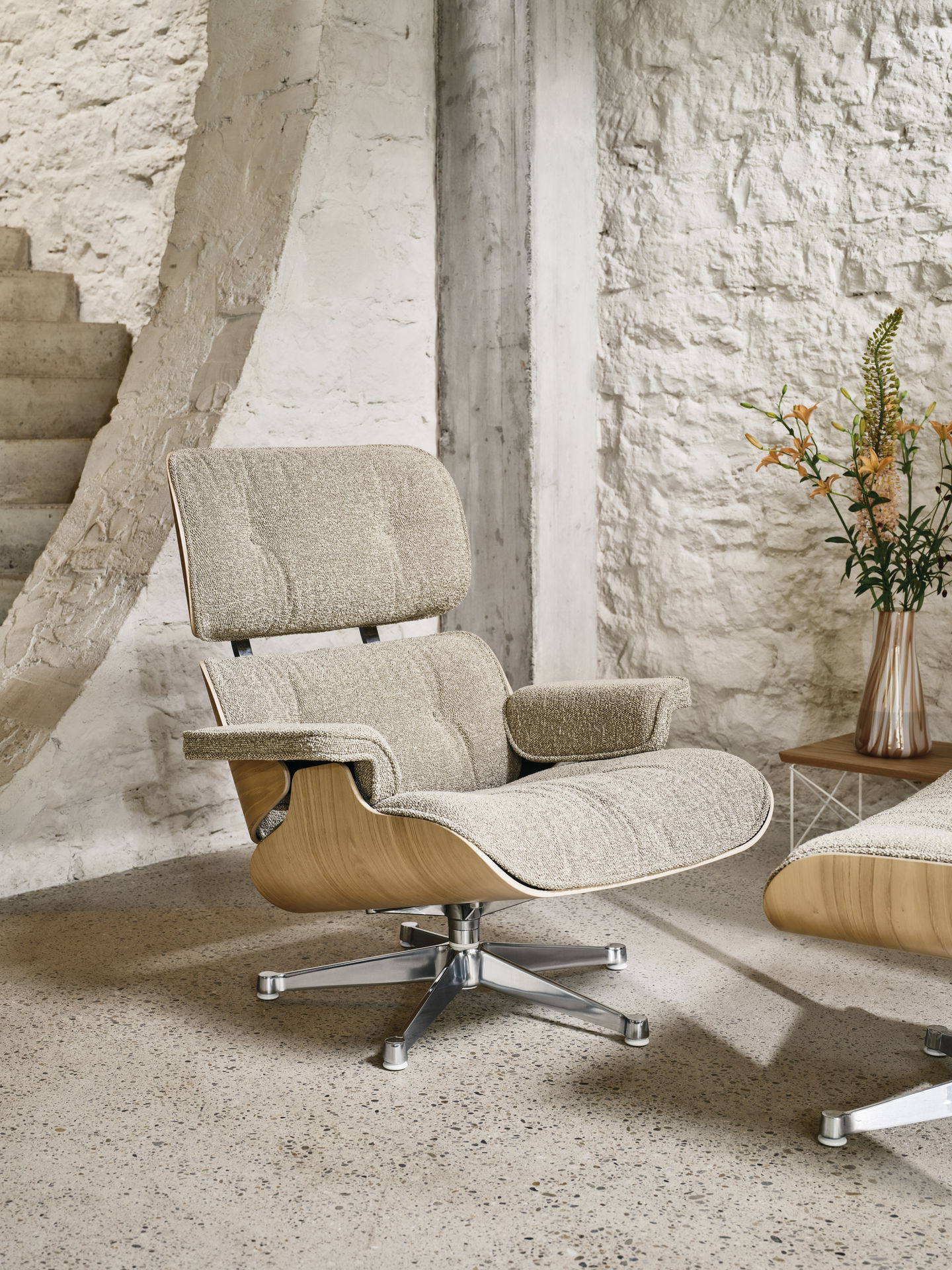 Beige Eames Lounge Chair Sessel mit Ottoman und Holzschale vor einer weißen Steinwand.