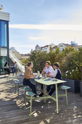 Drei Personen sitzen am grünen Bolder 3-Sitzer-Tisch von Fermob im Freien auf einer Dachterrasse.