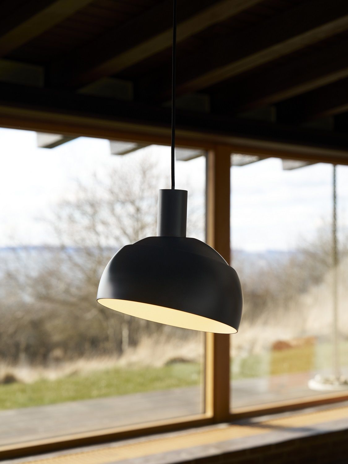 Schwarze FJ Elements Pendelleuchte von Louis Poulsen mit warmem Licht vor einem Fenster.