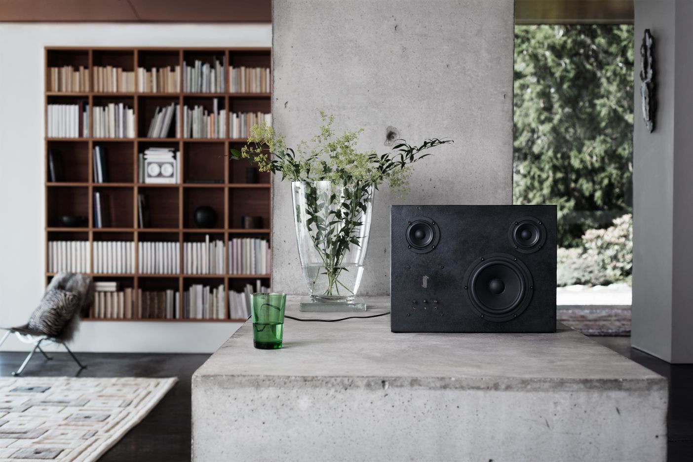 Schwarzer Stahl Lautsprecher auf Betonoberfläche mit Glasvase und Bücherregal im Hintergrund. Hi-Fi Lautsprecher für Musikliebhaber.
