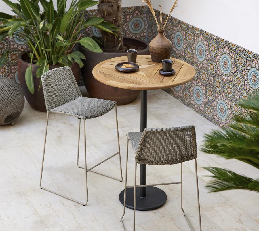 Zwei Cane-Line Breeze Barhocker in Taupe stehen an einem runden Gartentisch.