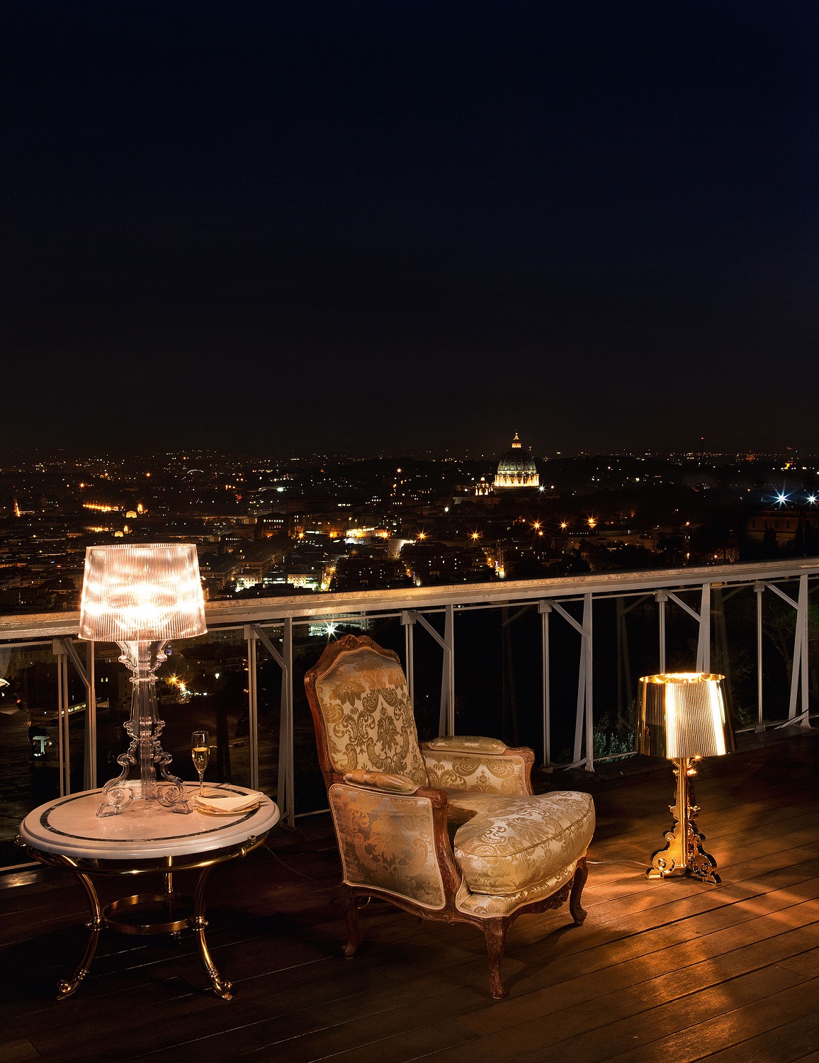 Bourgie Tischleuchte von Kartell, glasklar und dimmbar, auf einem Tisch mit Blick auf eine beleuchtete Stadt.