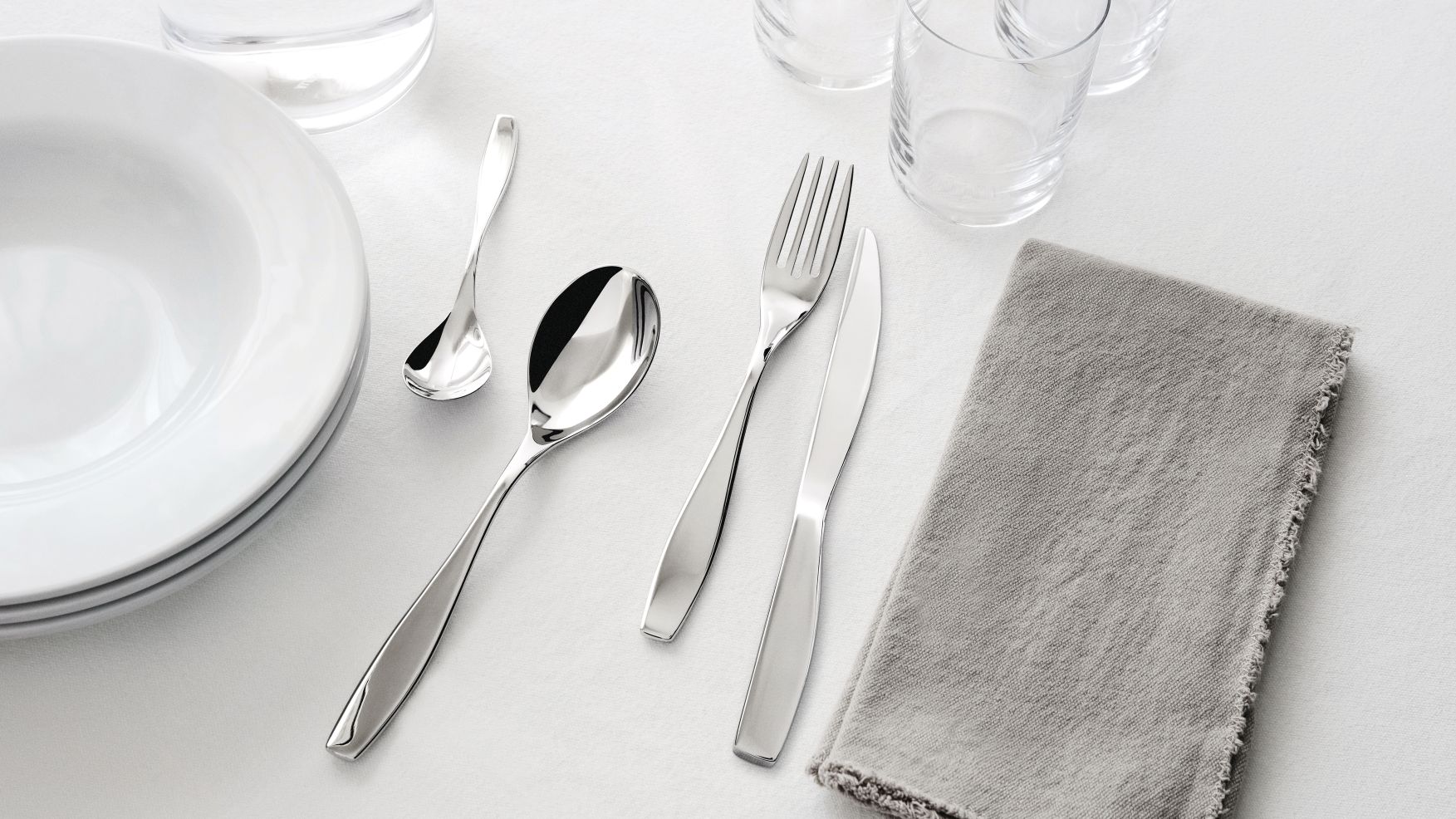 Nahaufnahme der Alessi Duna Besteckgarnitur, bestehend aus Messer, Gabel, Löffel und Dessertlöffel, neben Tellern und Gläsern.