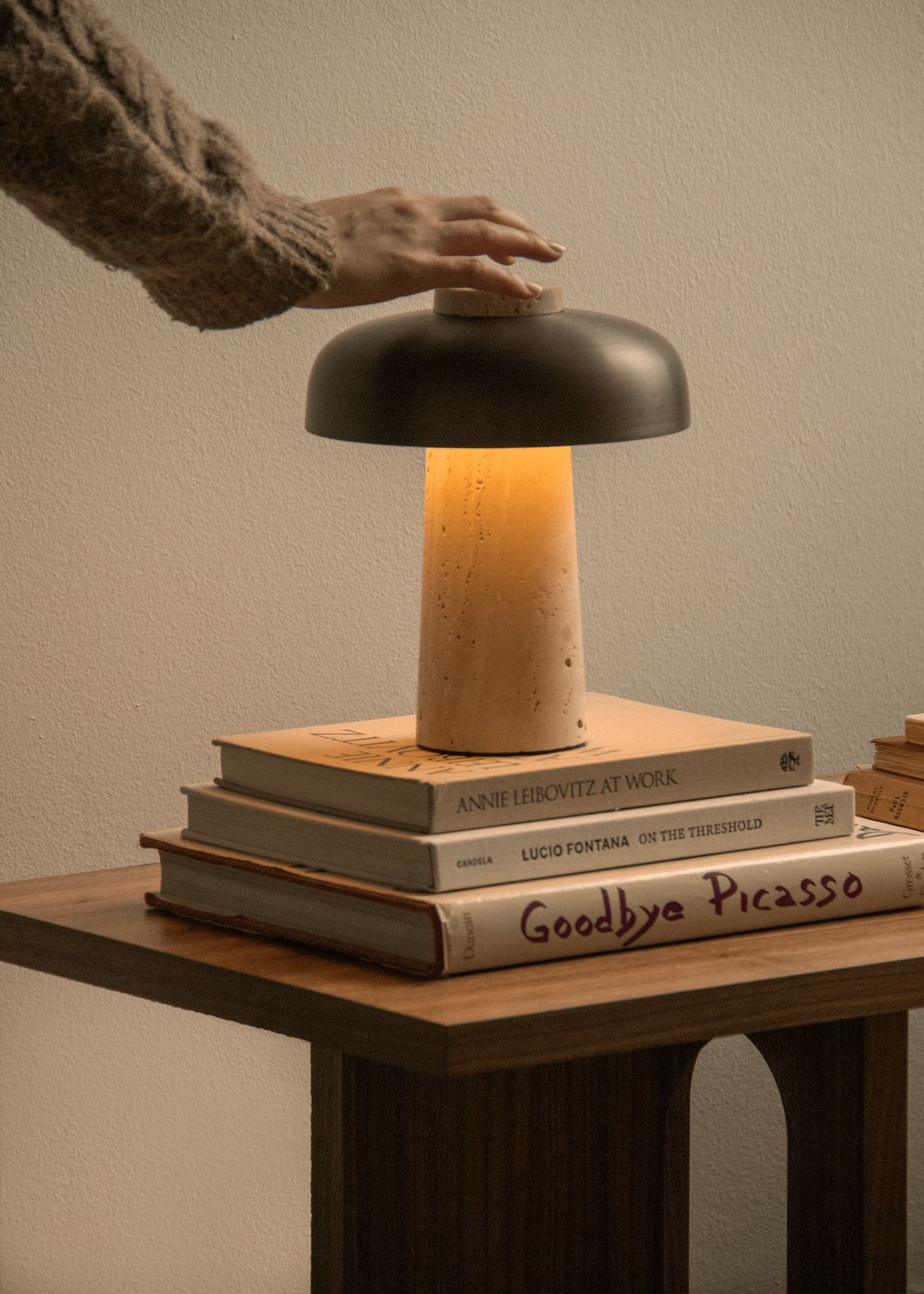 Audo Copenhagen Reverse Akkuleuchte auf Bücherstapel, Hand schaltet Lampe ein, warmes Licht.