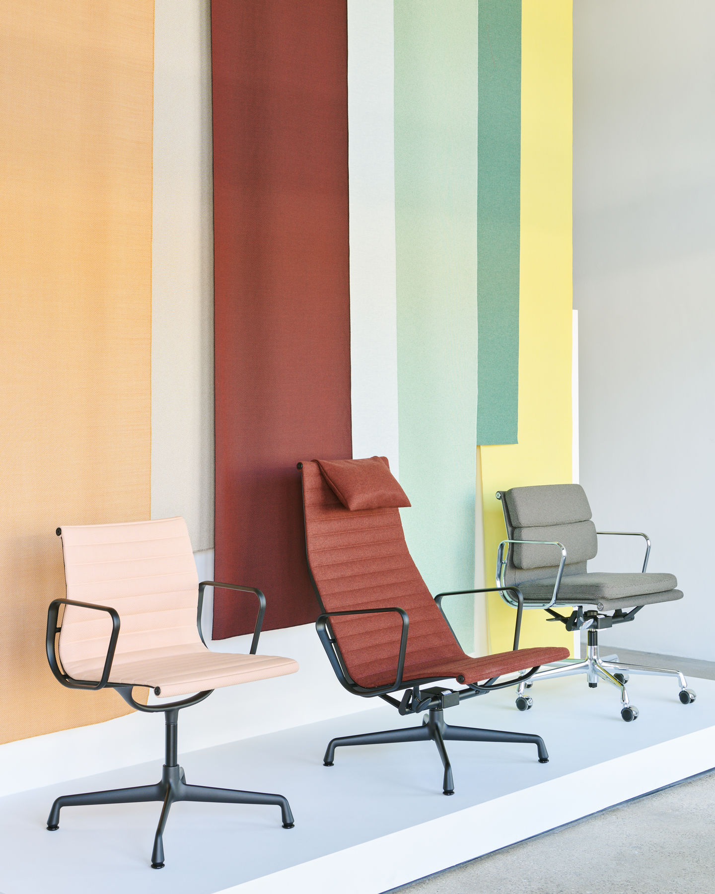 Drei Vitra Aluminium Chairs EA124 in verschiedenen Farben vor farbigem Hintergrund, Bürostühle mit Armlehnen.
