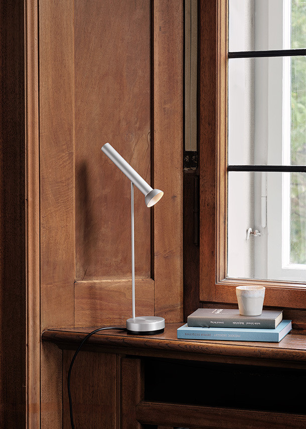 TOPOLED T Tischleuchte von Baltensweiler in Silber vor einem Fenster und einer Holzwand.