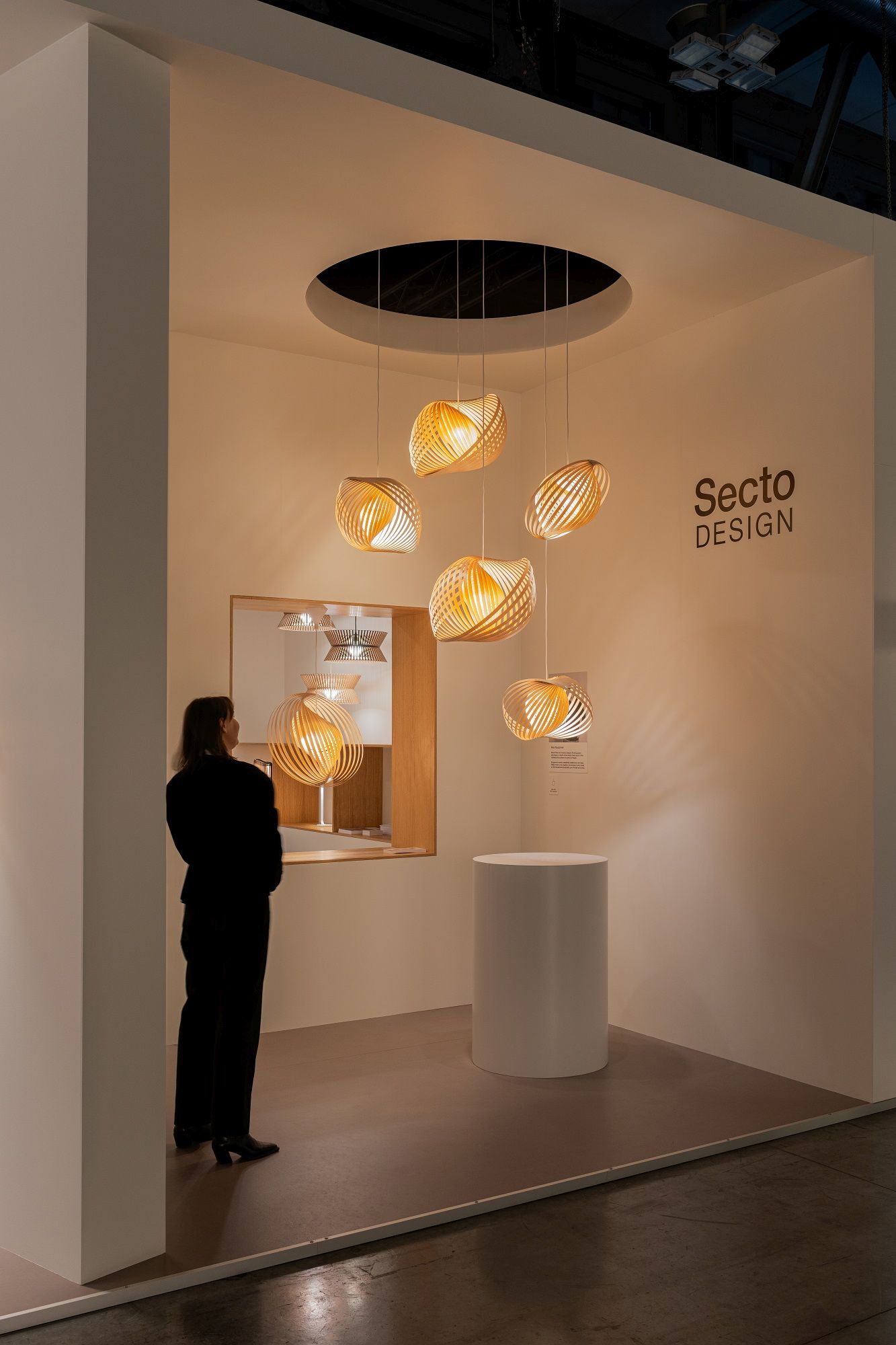 Adilo Pendelleuchte von Secto Design: Moderne Holzleuchten als Pendelleuchten-Ensemble in einem Ausstellungsraum mit Besucherin.
