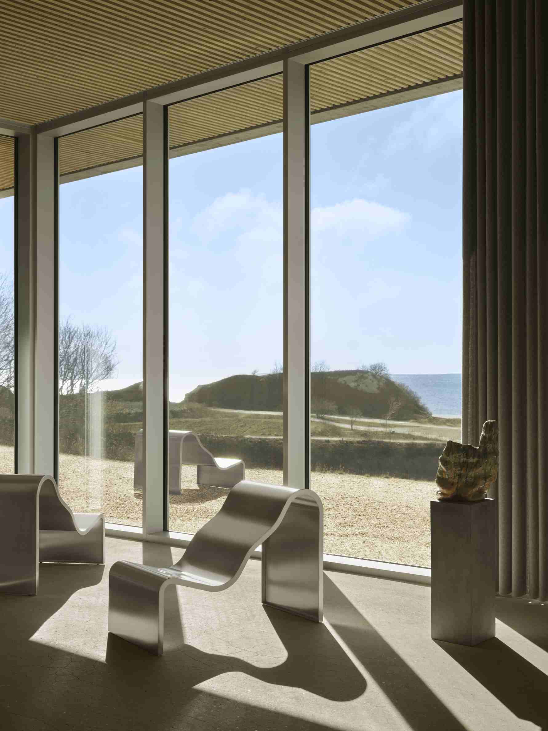 Muuto Dream Bench Indoor/Outdoor Bank vor Fenster mit Blick auf Dünenlandschaft und Meer.