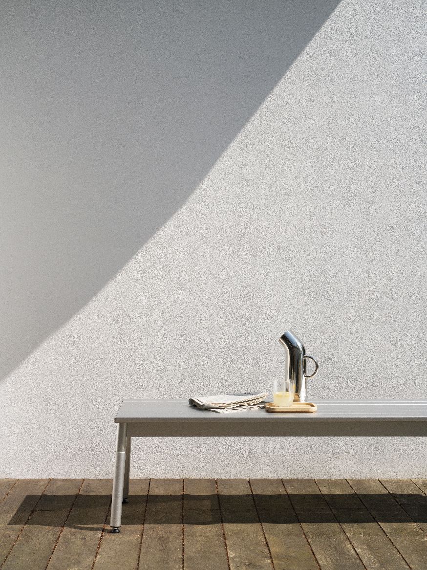 Normann Copenhagen Ex Bank Outdoor mit Getränkekrug und Glas auf Holzterrasse vor heller Wand.