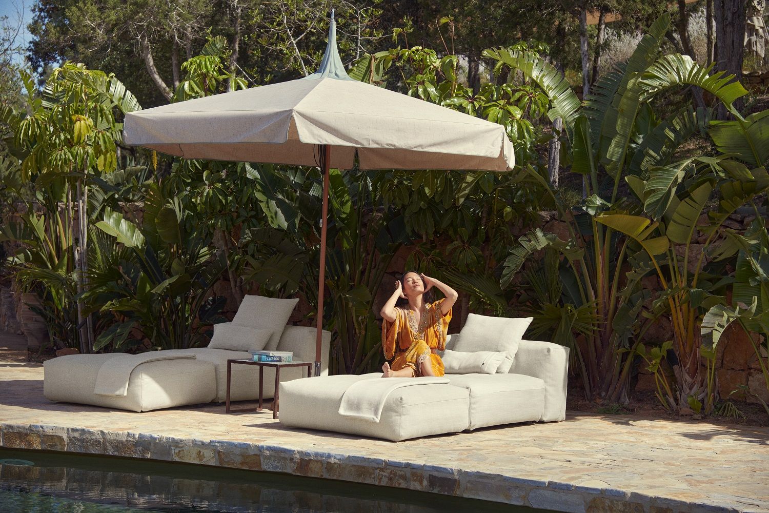 Frau entspannt auf einem beigen vetsak Noodle Pillow Pique Outdoor Kissen am Pool unter einem Sonnenschirm.