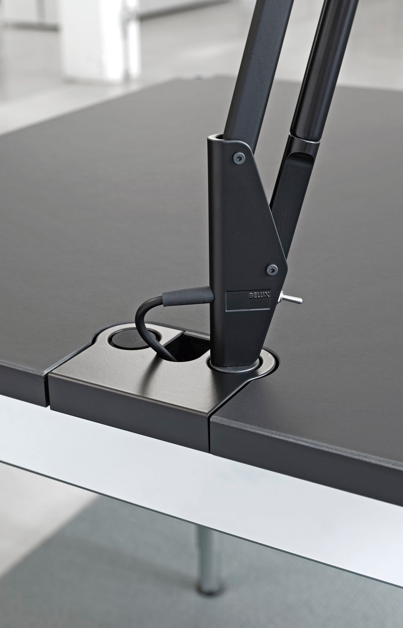 Detailaufnahme des Belux Lifto/Liftolino Tischleuchten Adapters USM Plus, montiert an einem schwarzen Bürotisch.