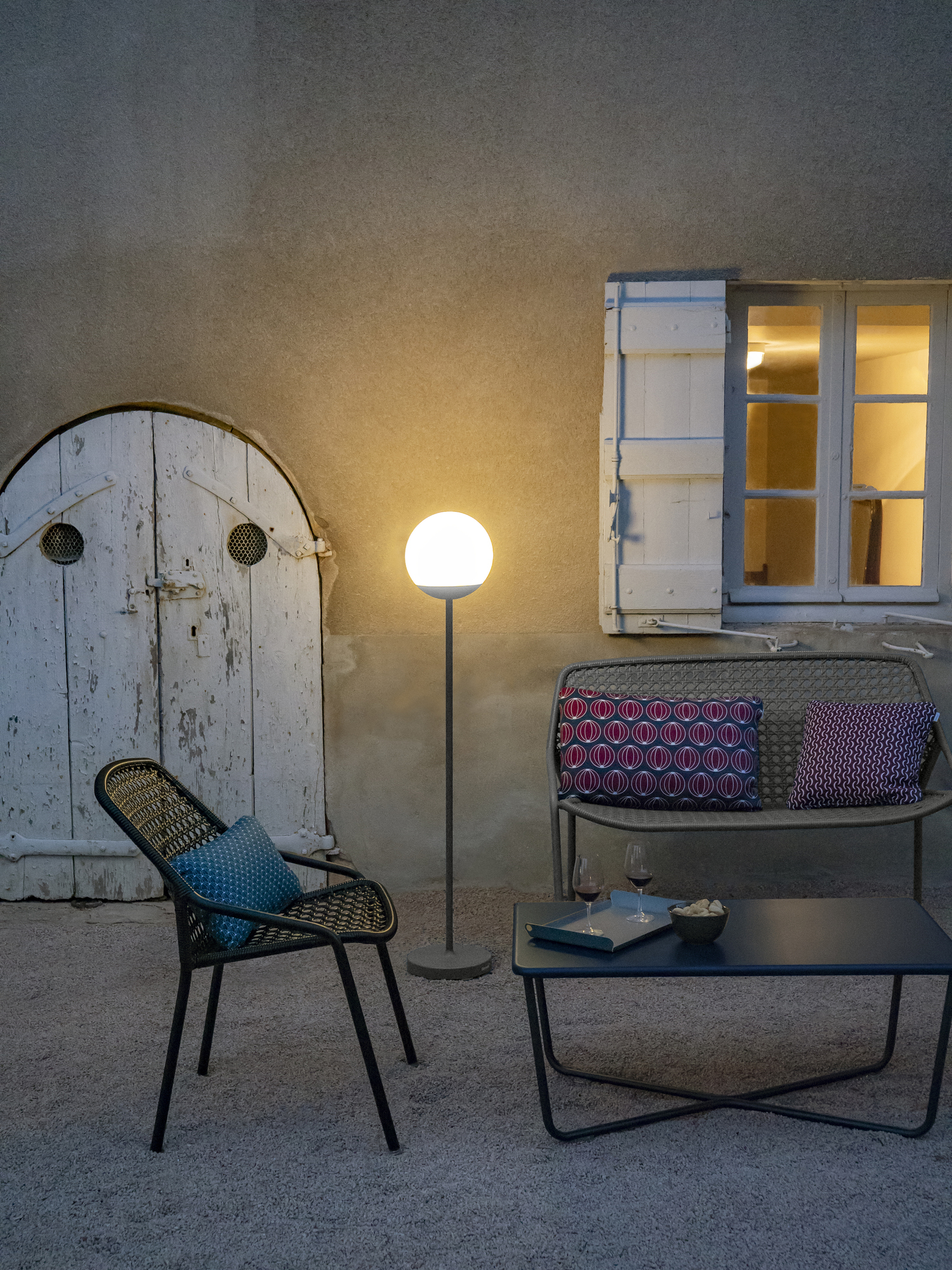 Anthrazitfarbene Mooon! Stehleuchte von Fermob beleuchtet eine gemütliche Outdoor-Sitzecke mit Tisch, Stuhl und Bank.