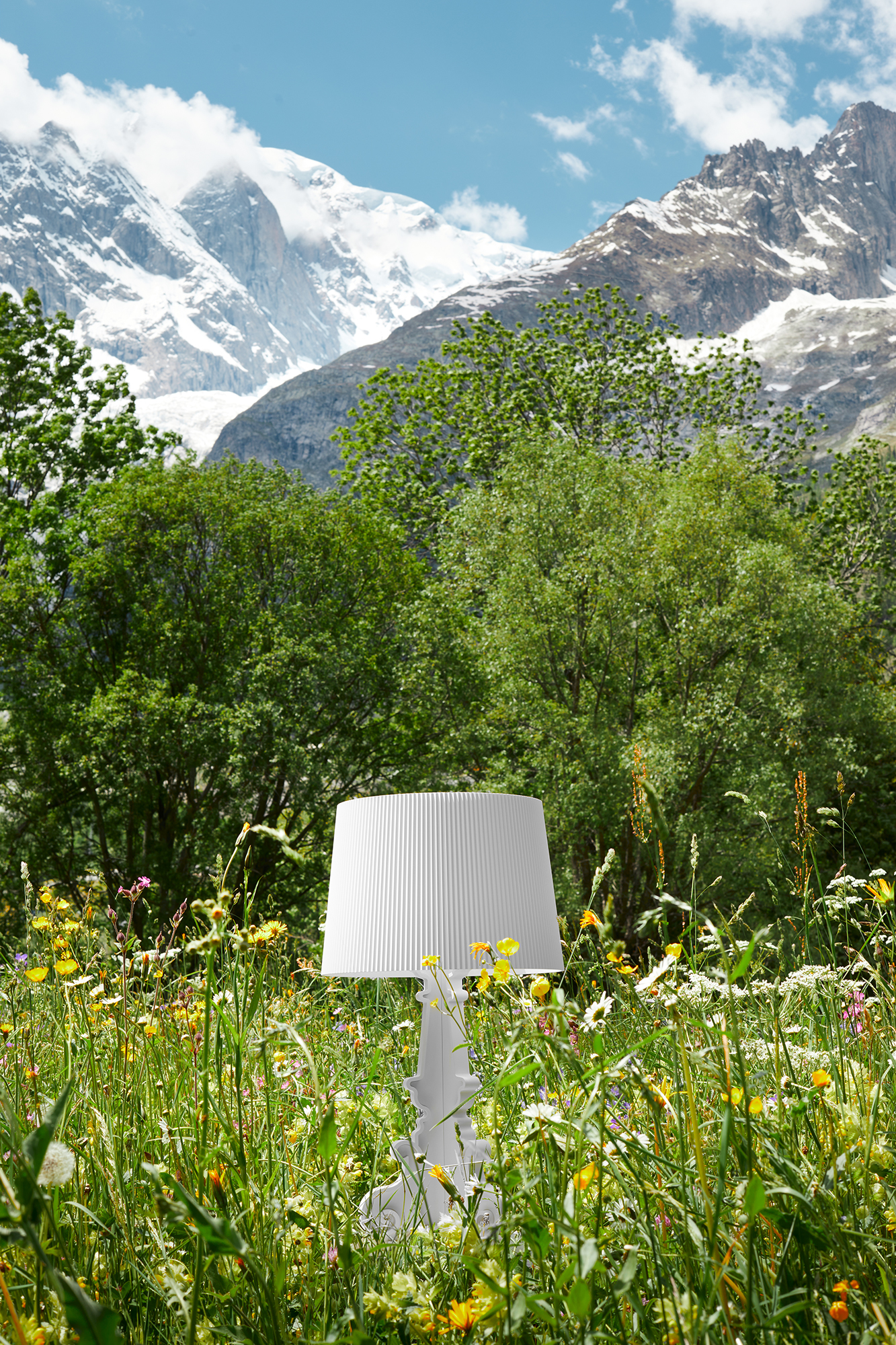 Weiße Kartell Bourgie Tischleuchte im Freien, umgeben von Blumenwiese und Berglandschaft.