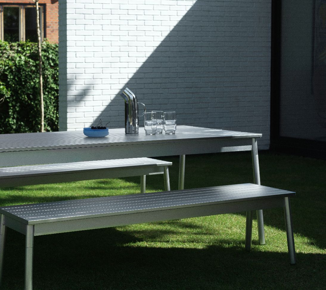 Normann Copenhagen Ex Tisch Outdoor mit Bänken auf Rasen, Geschirr und Getränke auf dem Gartentisch.