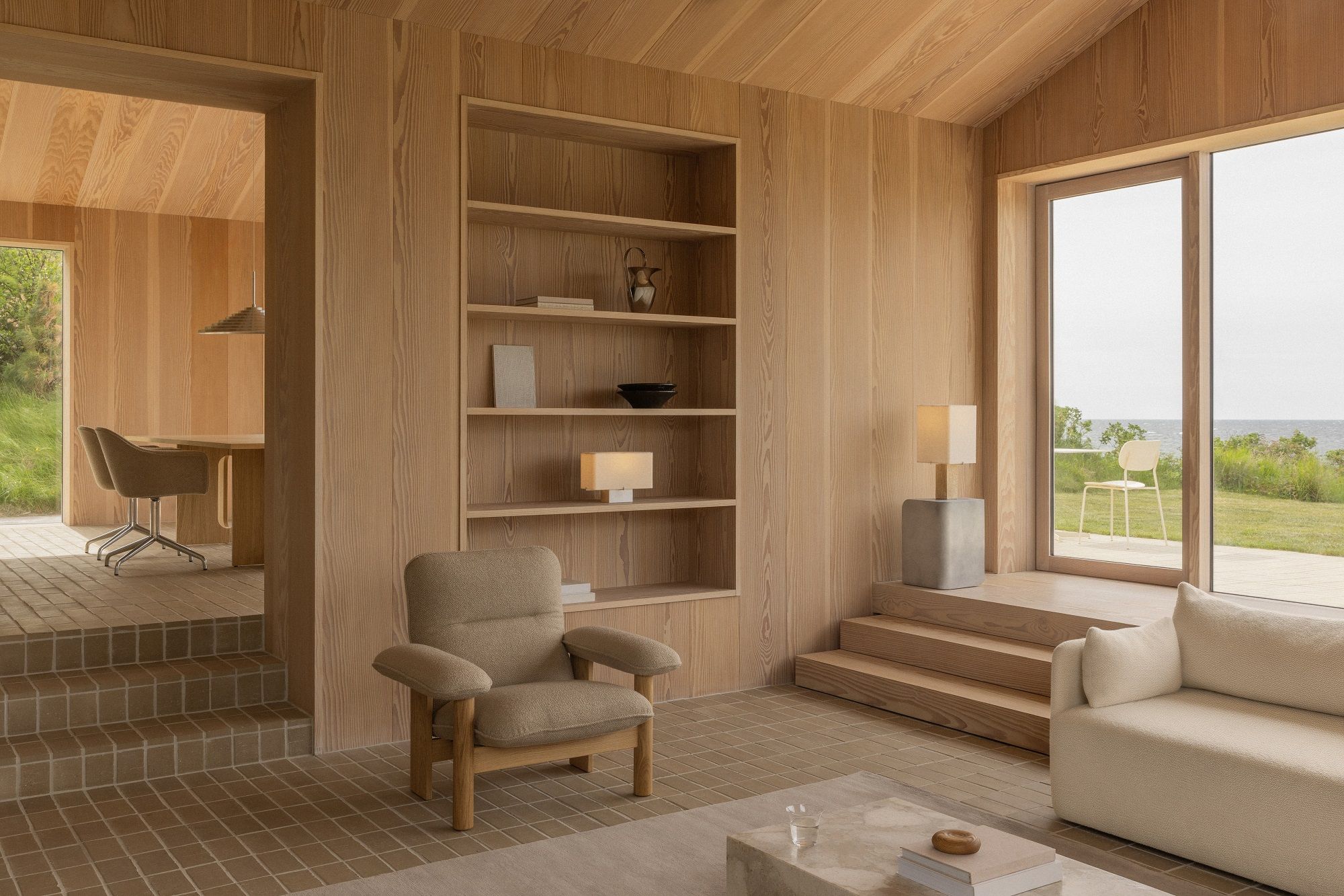 Helles Wohnzimmer mit Audo Copenhagen Offset Sofa, Sessel, Holzregal und Blick auf die Natur.