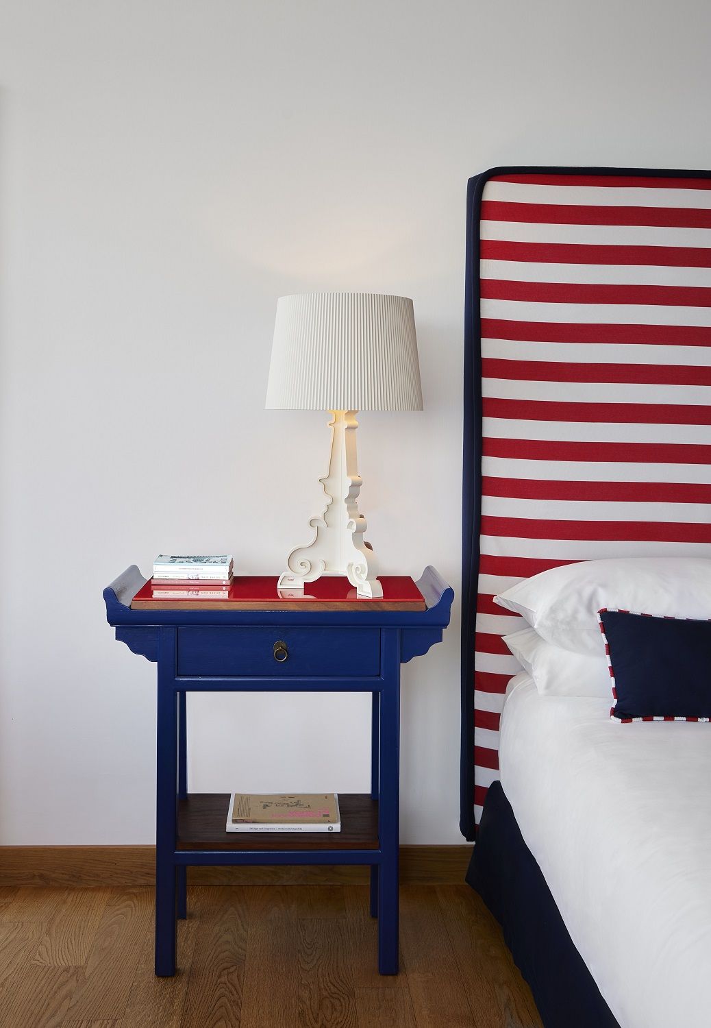 Weiße Bourgie Tischleuchte von Kartell auf blauem Nachttisch neben einem rot-weiß gestreiften Bett.