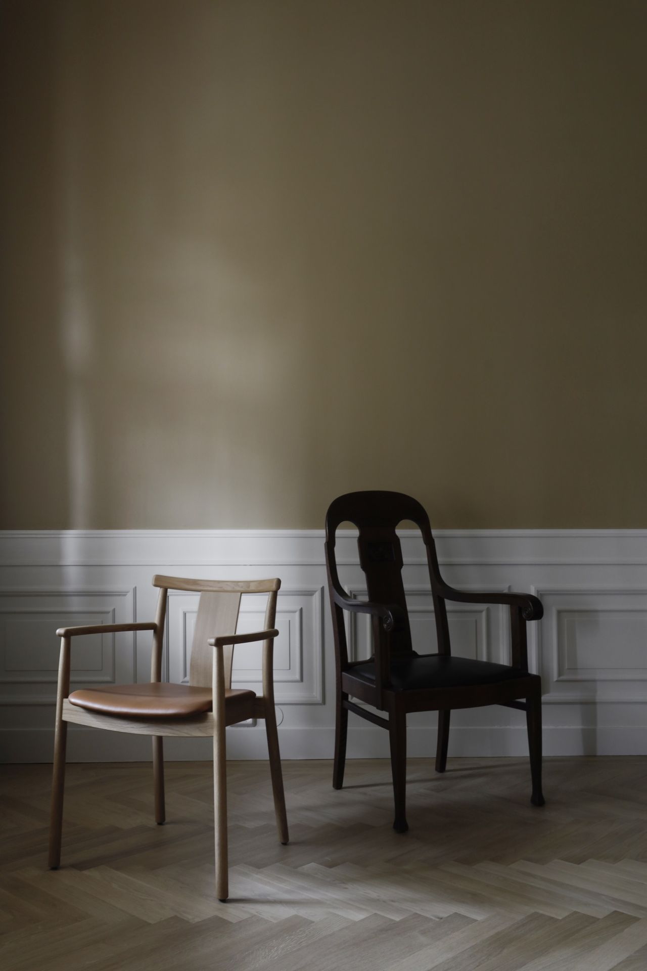 Zwei Esszimmerstühle: Heller Merkur Dining Chair mit Armlehnen und dunkler Holzstuhl vor Wandpaneelen.