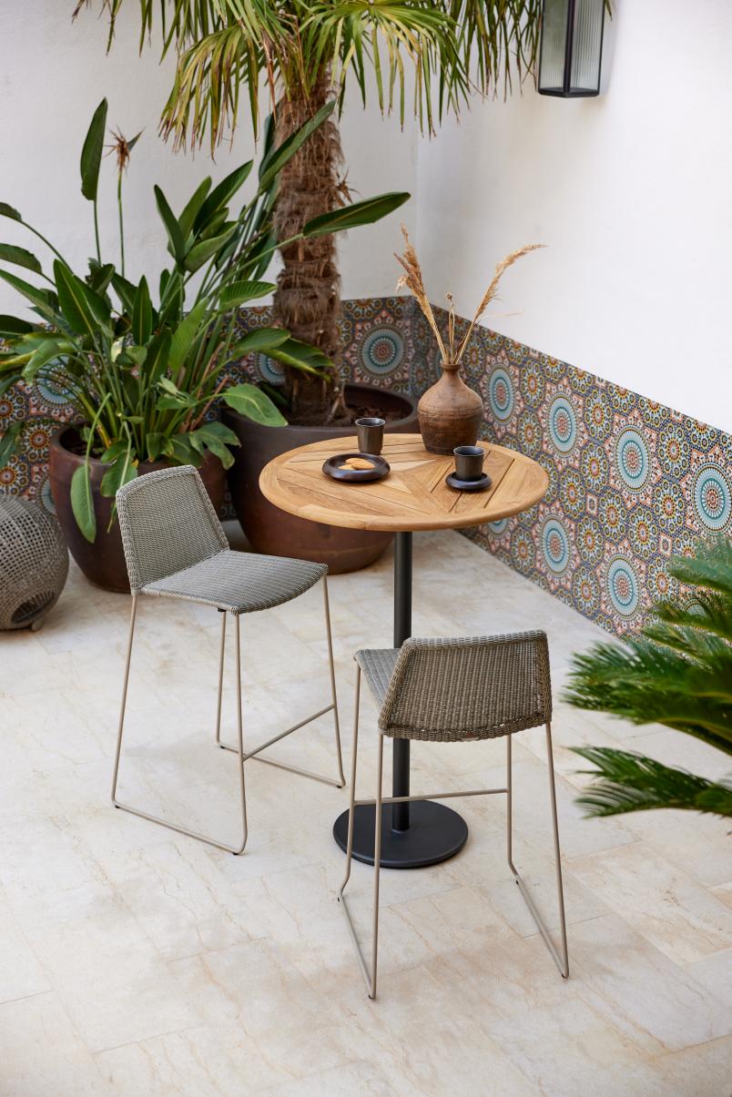 Zwei Cane-Line Barhocker in Taupe mit Outdoor-Tisch auf einer Terrasse mit Pflanzen.
