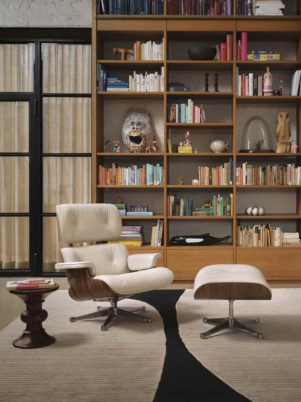 Beiger Eames Lounge Chair Sessel mit Hocker vor Bücherregal, Designklassiker für Wohnzimmer und Büro.