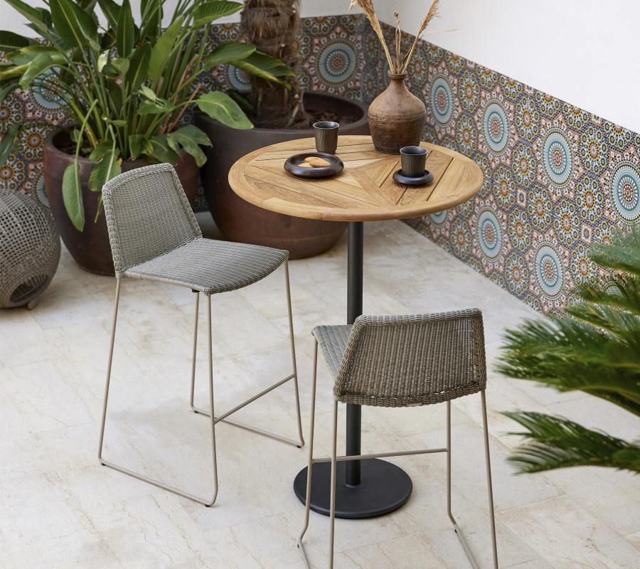 Zwei Cane-Line Breeze Barhocker in Taupe stehen an einem runden Gartentisch.