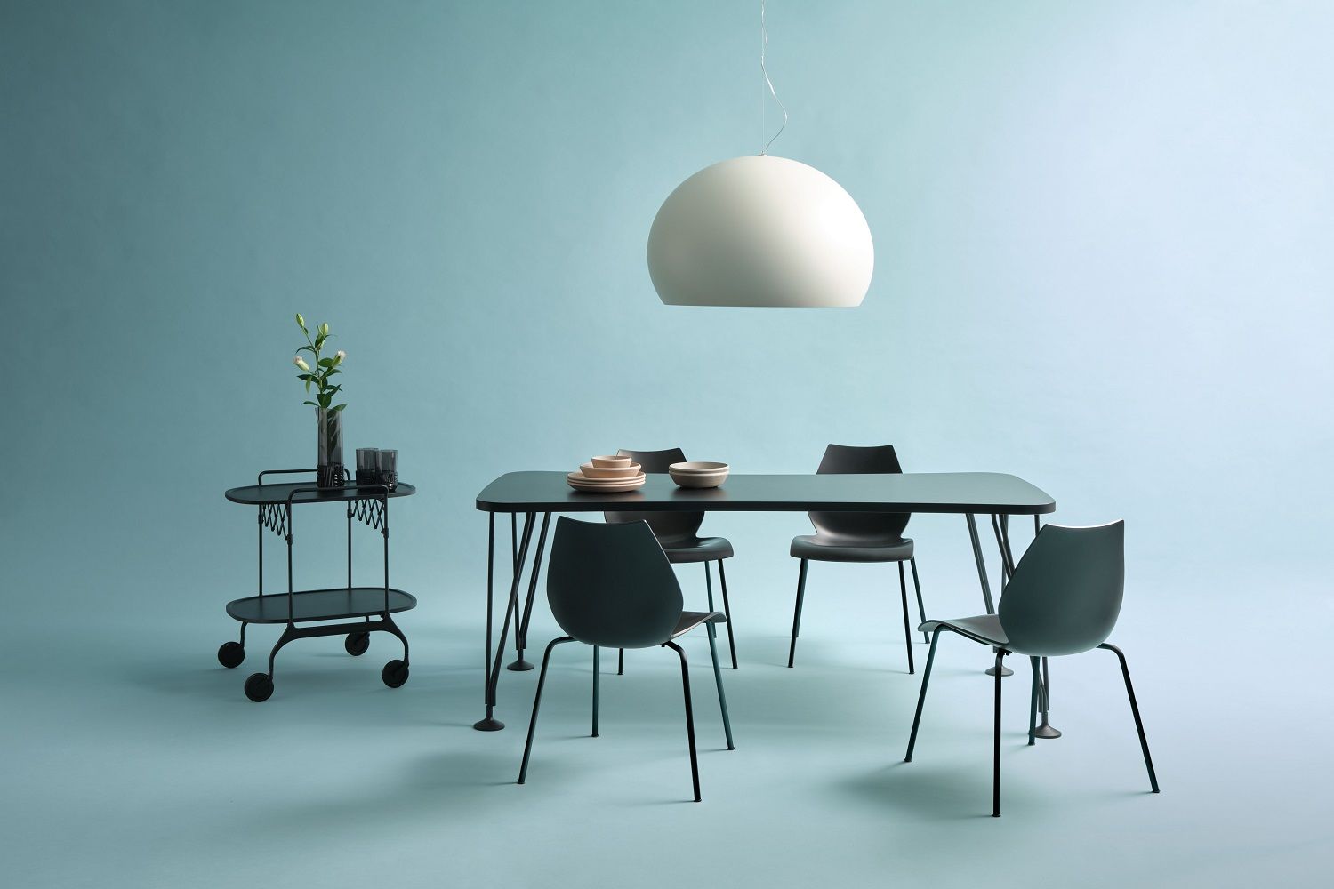 Helles Esszimmer mit Kartell Fly Pendelleuchte, schwarzem Tisch, Stühlen und Servierwagen vor blauem Hintergrund.