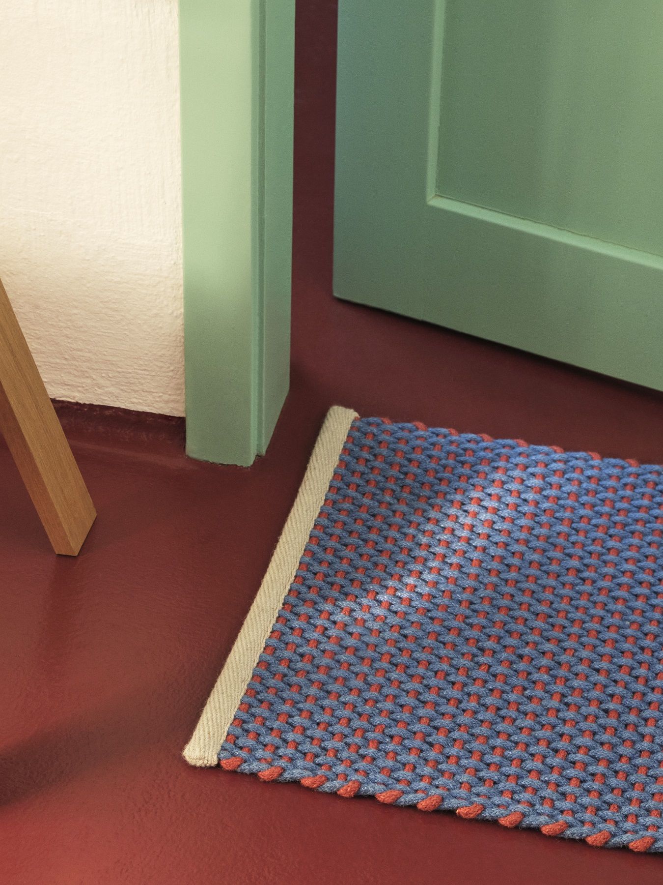 Hay Teppich Fußmatte aus Baumwolle, blau und rot gewebt, vor einer grünen Tür auf rotem Boden.