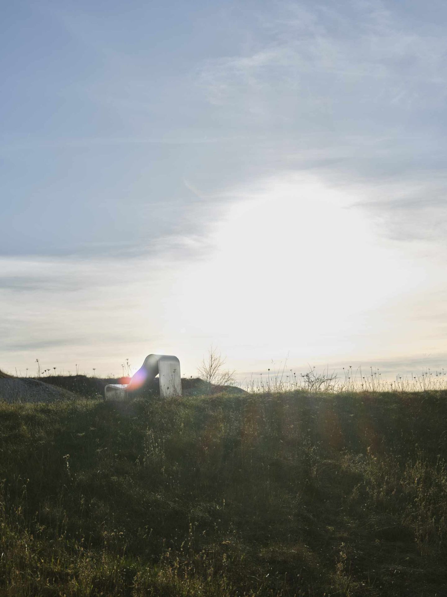 Muuto Dream Bench Outdoor Bank auf einer Wiese mit Gras und hellem Sonnenlicht im Hintergrund.