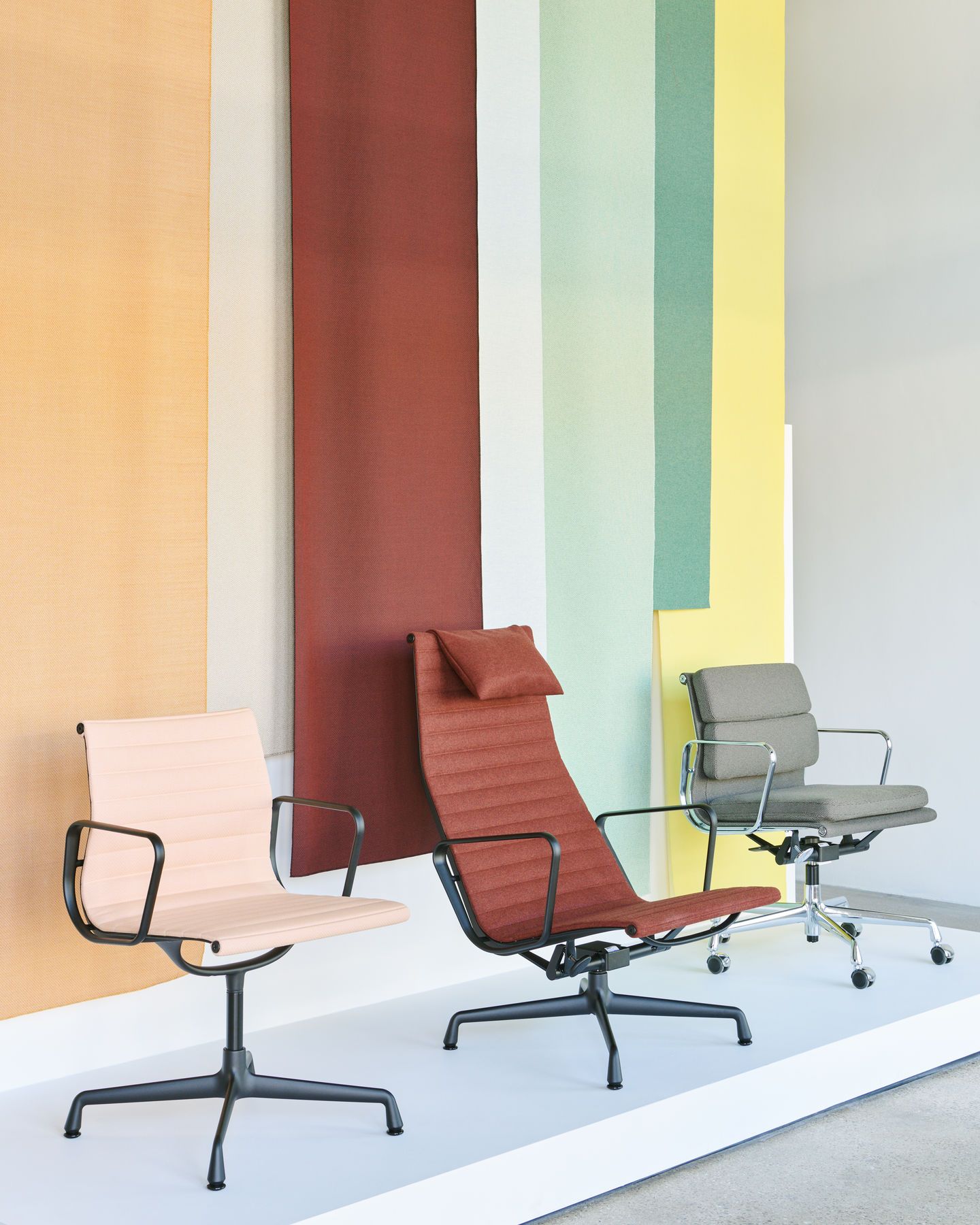 Drei Vitra Aluminium Chairs EA124 in verschiedenen Farben vor farbigem Hintergrund, Bürostühle mit Armlehnen.