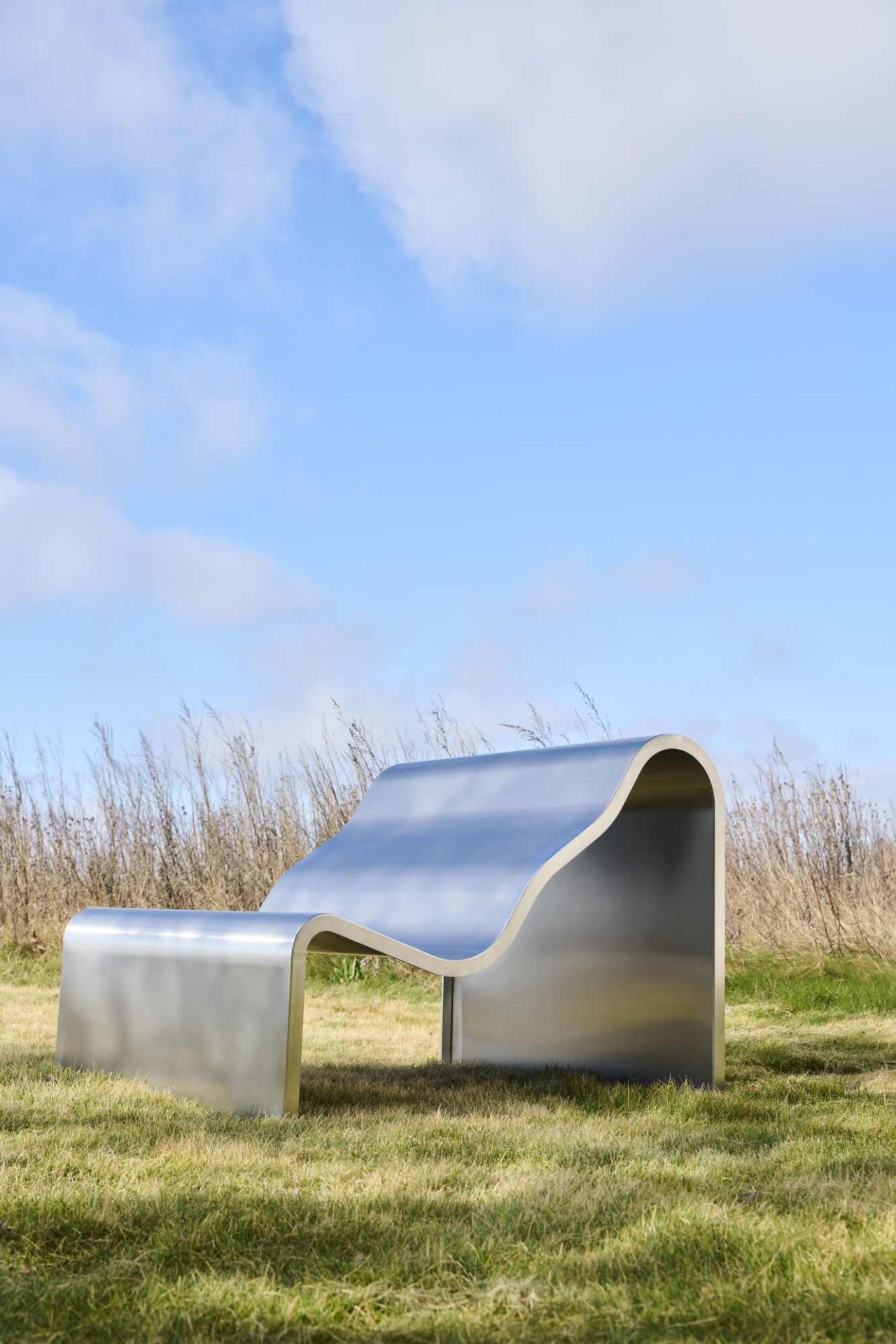 Muuto Dream Bench Indoor/Outdoor Bank aus gebogenem Edelstahl auf einer grünen Wiese unter blauem Himmel.