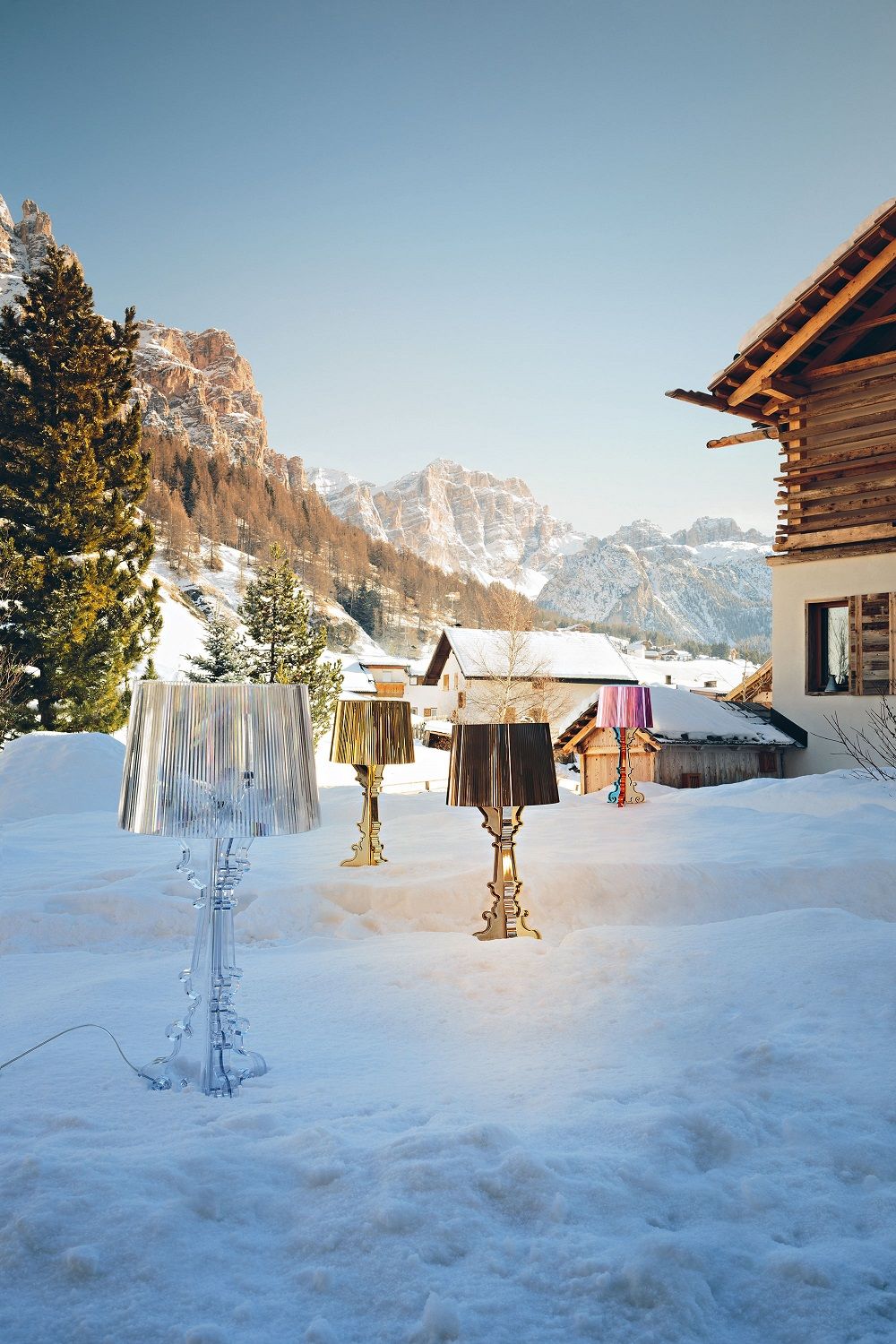 Mehrfarbige Bourgie Tischleuchten von Kartell im Schnee vor verschneiter Bergkulisse und Holzhäusern.