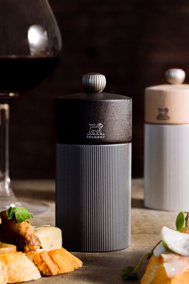 Peugeot Line Pfeffermühle H 12 cm Graphit & Carbon neben Speisen und einem Glas Rotwein.
