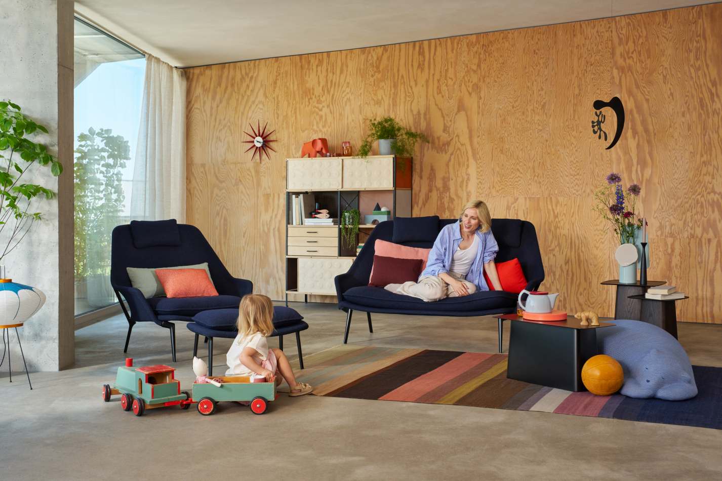 Wohnzimmer mit blauen Vitra Slow Chair Sessel und Ottoman, Frau sitzt auf Sofa.