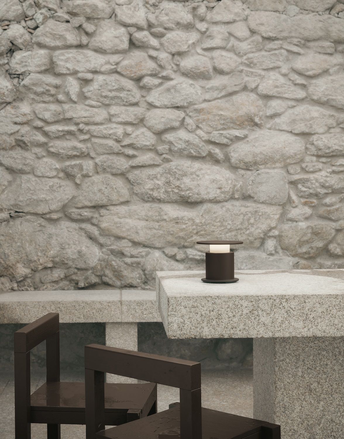 w241 Faro Tischleuchte von wästberg auf Steintisch mit Stühlen vor Steinwand. Moderne Akkuleuchte.