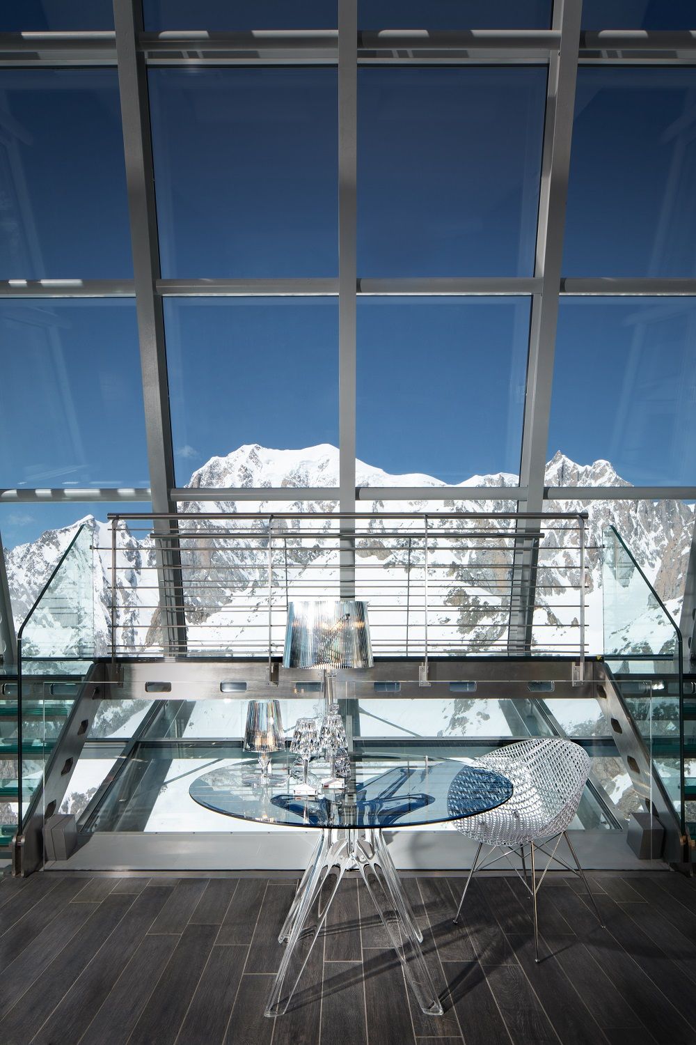Bourgie Tischleuchte silber von Kartell auf gläsernem Tisch mit Bergpanorama im Hintergrund. Moderne Designleuchte.