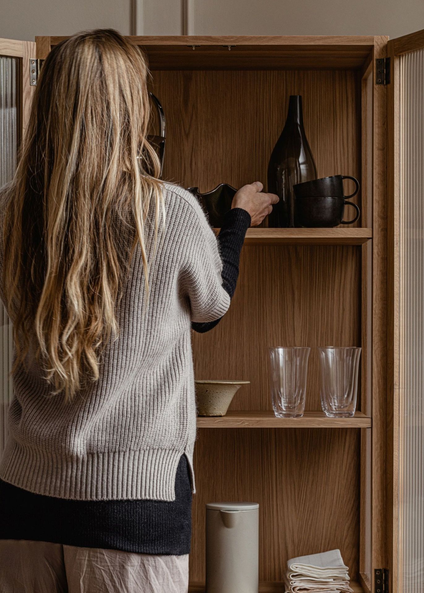 Frau räumt Geschirr in einen Curiosity Cabinet Schrank von Audo Copenhagen aus hellem Holz ein.