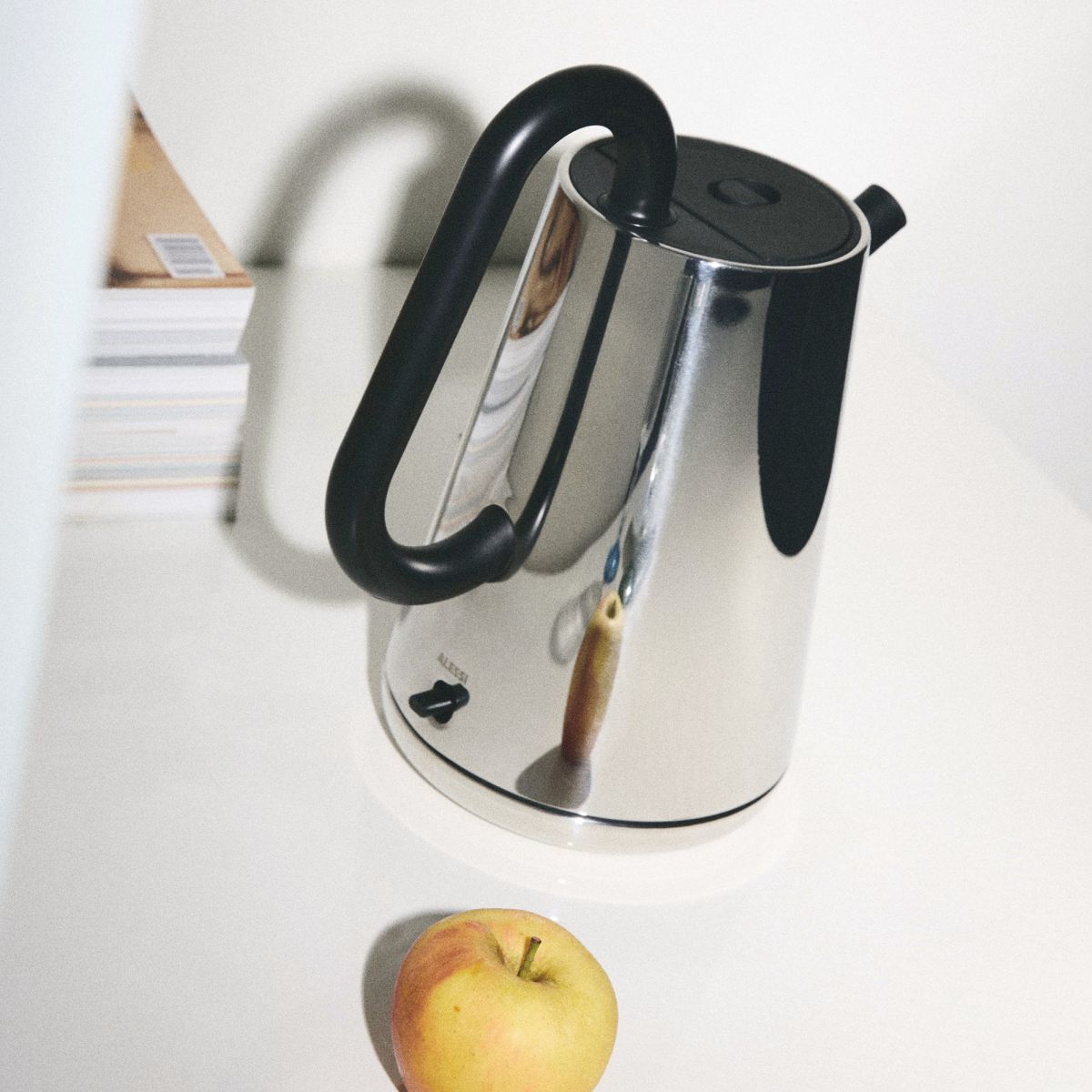 Alessi Toru Wasserkocher aus Edelstahl mit schwarzem Griff und Apfel auf weißem Tisch.