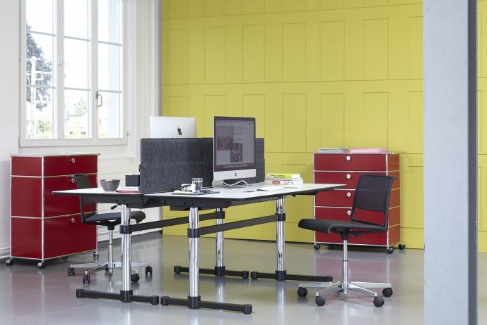 USM Haller Büroeinrichtung mit Schreibtisch, Privacy Panels, Bürostuhl und roten Sideboards vor gelber Wand.