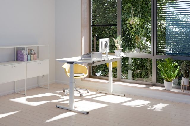 Heller Büroraum mit Palmberg PiD Sitz-Stehtisch, Monitor, gelbem Bürostuhl und Fenster mit Blick ins Grüne.