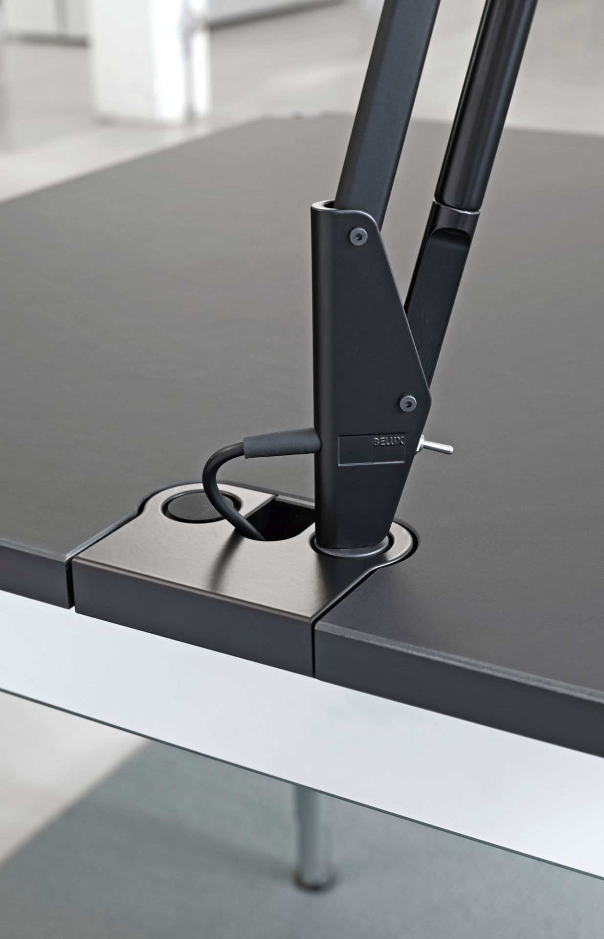 Detailaufnahme des Belux Lifto/Liftolino Tischleuchten Adapters USM Plus, montiert an einem schwarzen Bürotisch.