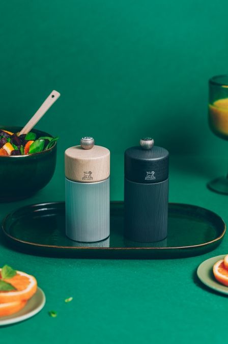 Zwei Peugeot Line Pfeffermühlen in Graphit und Carbon auf einem grünen Tisch mit Speisen.